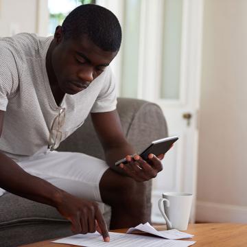 Un hombre sentado en un sofá con una computadora portátil y una taza de café, absorto en su trabajo y disfrutando de una bebida caliente.